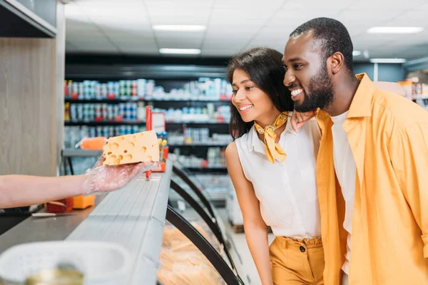 Cropped image of shop assistant giving cheese to smiling african american couple in supermarket — Stock Photo