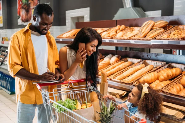 Homem americano africano marcação lista de compras, enquanto sua esposa falando no smartphone e olhando para a filha no supermercado — Fotografia de Stock