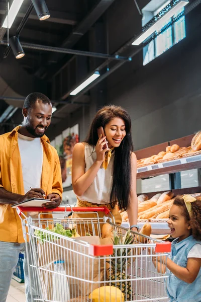 Усміхнена афроамериканська жінка розмовляє на смартфоні, а її чоловік і дочка стоять поруч із торговим візком у супермаркеті — стокове фото