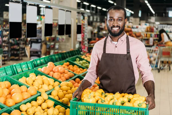 Heureux afro-américain homme assistant de magasin dans tablier tenant boîte avec poivrons dans l'épicerie — Photo de stock