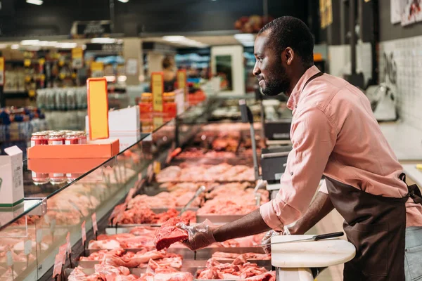 Vue latérale du boucher mâle afro-américain dans un tablier donnant un steak de viande crue dans un supermarché — Photo de stock