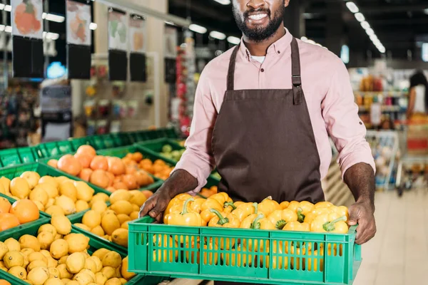 Cropped image of african american male shop assistant in apron holding box with bell peppers in grocery store — Stock Photo