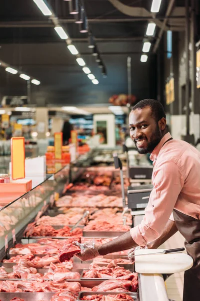 Carnicero masculino afroamericano en delantal sosteniendo filete de carne cruda y mirando a la cámara en el supermercado - foto de stock