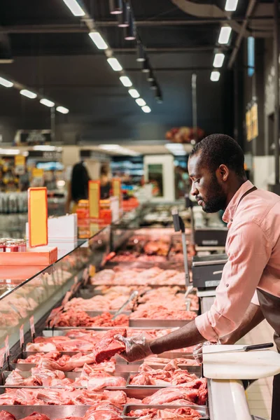 Focalizzato macello afroamericano maschio in grembiule prendendo bistecca di carne cruda nel supermercato — Foto stock