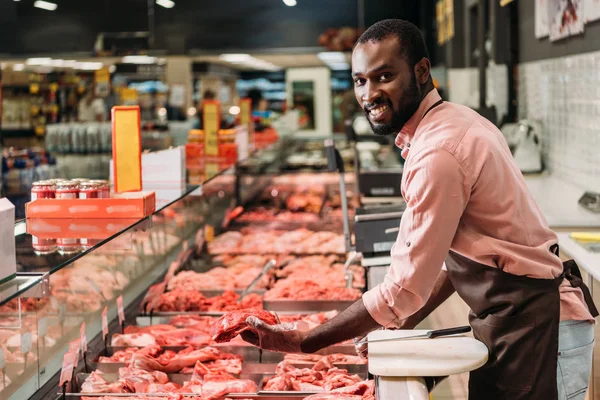 Enfoque selectivo del carnicero afroamericano en delantal con filete de carne cruda en la tienda de comestibles - foto de stock