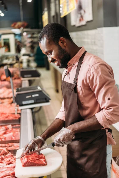 Sorrindo afro-americano assistente de loja masculino em avental cortando bife de carne crua no supermercado — Fotografia de Stock