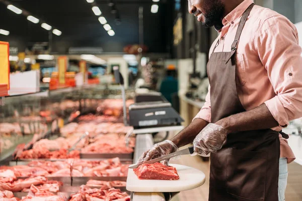 Cropped image of african american male shop assistant in apron cutting raw meat in grocery store — Stock Photo