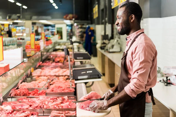 Vista lateral del carnicero afroamericano sonriente en delantal cortando carne cruda en la tienda de comestibles - foto de stock