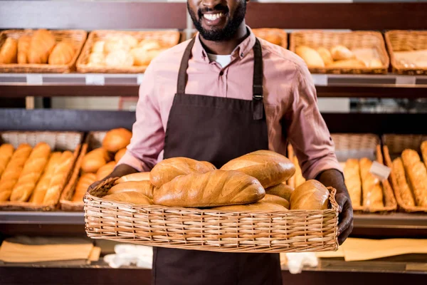 Cropped image of happy african american male shop assistant in apron holding loaves of bread in hypermarket — Stock Photo