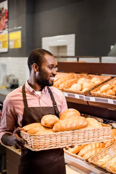 Africano americano tienda asistente en delantal celebración de panes en el supermercado - foto de stock