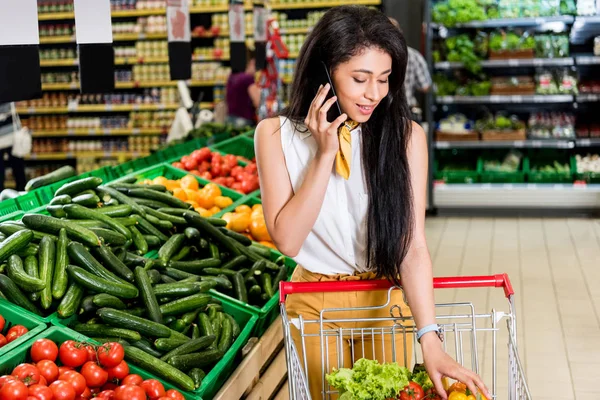 Young african american woman talking on smartphone near shopping trolley in grocery store — Stock Photo