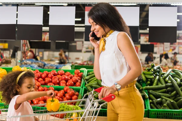 Mujer afroamericana hablando por teléfono inteligente mientras su hija está sentada en el carrito de compras en el supermercado - foto de stock