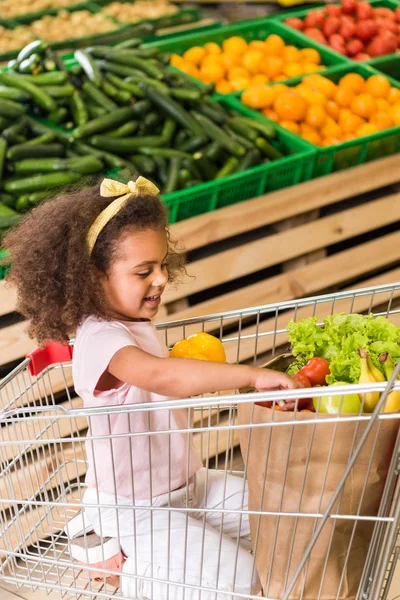 Niño afroamericano feliz sentado en el carro de la compra en la tienda de comestibles - foto de stock