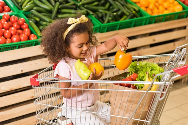 Niño afroamericano sonriente poniendo pimientos en bolsa de papel mientras está sentado en el carrito de la compra en el supermercado - foto de stock