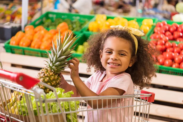 Bambino afroamericano sorridente seduto nel carrello della spesa con ananas nel negozio di alimentari — Foto stock