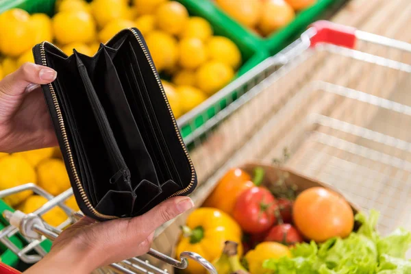 Imagen recortada de la mujer mostrando la cartera vacía cerca del carro de la compra en la tienda de comestibles - foto de stock