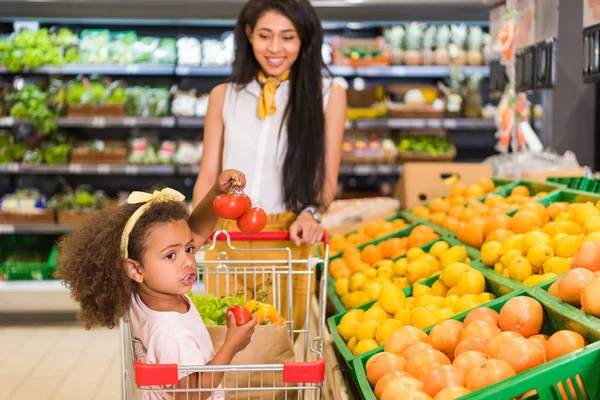 African american little child sitting in shopping trolley while her mother carrying it near fruit department in supermarket — Stock Photo