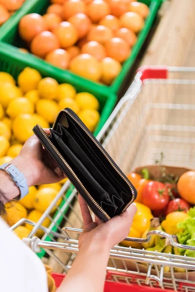Cropped image of woman showing empty purse near shopping trolley in supermarket — Stock Photo