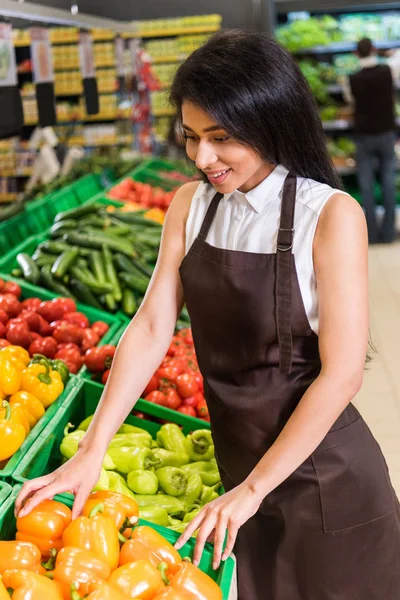 Sorrindo afro-americana assistente loja feminina em avental organizando pimentas no supermercado — Fotografia de Stock