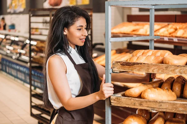 Lächelnd afrikanisch-amerikanische Verkäuferin in Schürze, die frisches Gebäck im Lebensmittelladen arrangiert — Stockfoto