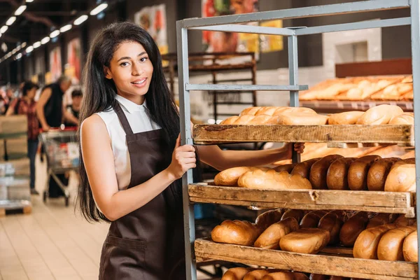 Young african american female shop assistant in apron arranging fresh pastry in supermarket — Stock Photo