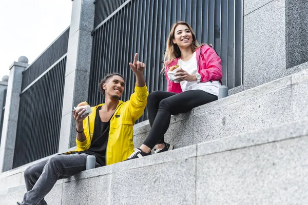 Young mixed race man with burger pointing by finger to asian girlfriend at city street — Stock Photo