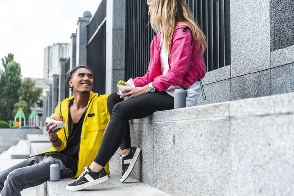 Enfoque selectivo de pareja sonriente comiendo hamburguesas y sentado cerca de bebidas gaseosas en la calle de la ciudad - foto de stock