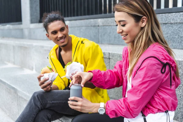 Smiling asian woman opening soda drink while her boyfriend holding burgers at urban street — Stock Photo