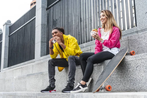 Joven pareja multicultural de patinadores comiendo hamburguesas en la calle de la ciudad - foto de stock