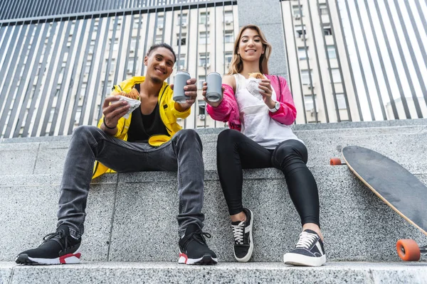 Vista basso angolo di sorridente coppia multietnica di skateboarder in possesso di hamburger e mostrando bevande gassate in strada città — Foto stock