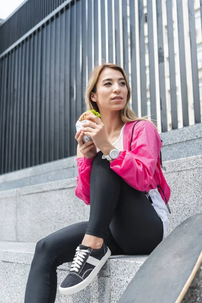 Attractive young asian woman holding burger near skateboard at urban street — Stock Photo