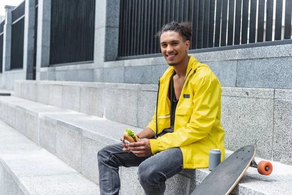 Jovem sorrindo misto raça patinador masculino sentado com hambúrguer e refrigerante perto de skate na rua da cidade — Fotografia de Stock