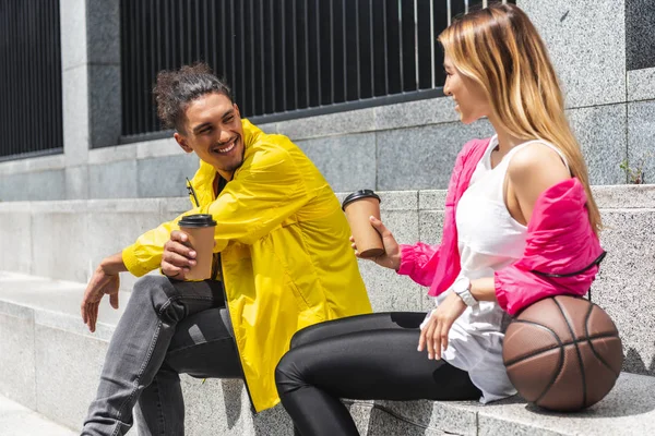 Sonriente pareja multicultural con pelota para baloncesto bebiendo café en la calle de la ciudad - foto de stock