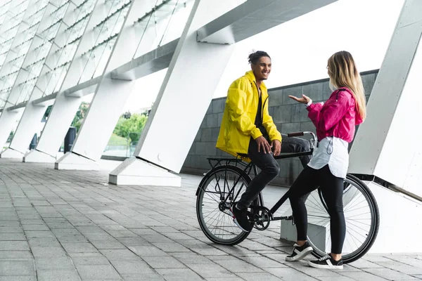 Sonriente hombre de raza mixta sentado en bicicleta y hablando con su novia en la calle - foto de stock