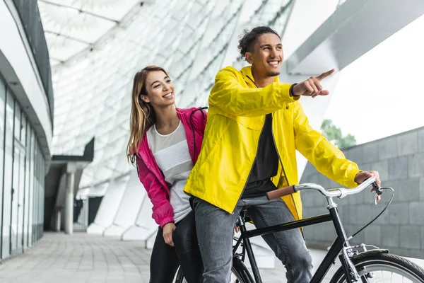 Sonriente hombre de raza mixta sentado en bicicleta y señalando con el dedo a su novia en la calle de la ciudad - foto de stock