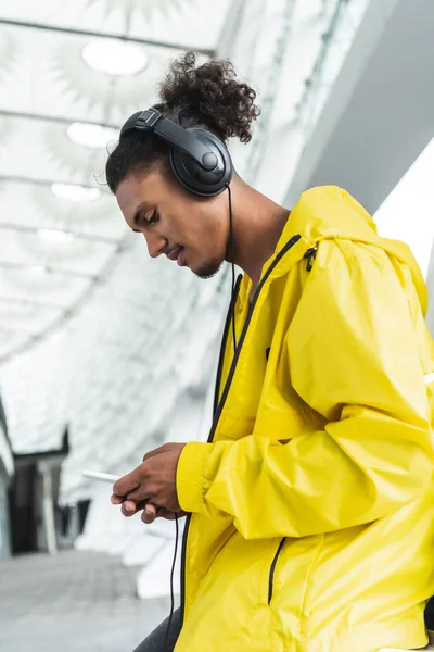Side view of smiling mixed race man in headphones listening music with smartphone at city street — Stock Photo