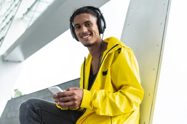 Smiling mixed race man in headphones listening music with smartphone and looking at camera at city street — Stock Photo