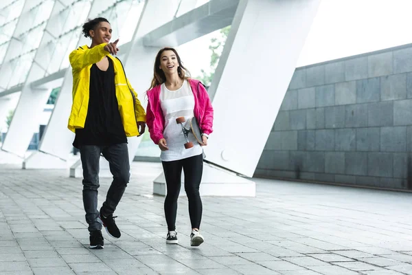 Hombre de raza mixta señalando con el dedo a la novia sonriente con monopatín en la calle - foto de stock