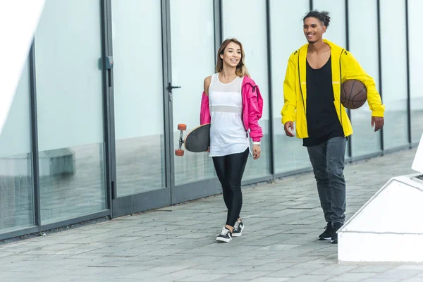 Happy multicultural couple walking with skateboard and basketball ball at city street — Stock Photo