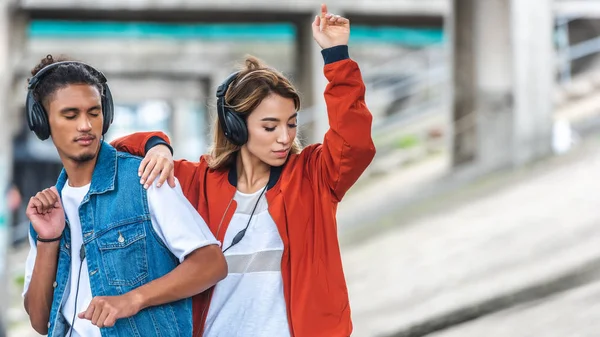 Joven pareja multicultural escuchando música con auriculares y bailando en la calle de la ciudad - foto de stock