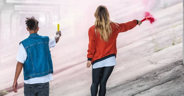 Rear view of stylish couple holding colorful smoke bombs at city street — Stock Photo