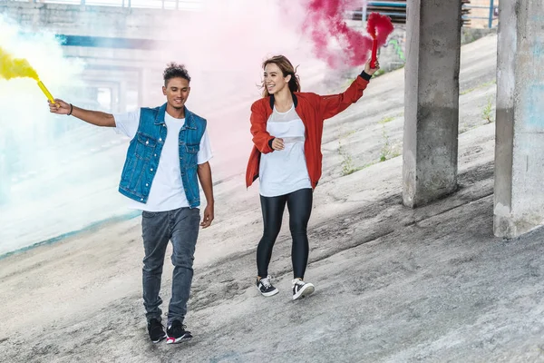 Smiling stylish couple holding colorful smoke bombs at city street — Stock Photo