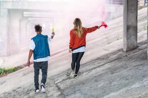 Rear view of stylish couple holding colorful smoke bombs at city street — Stock Photo