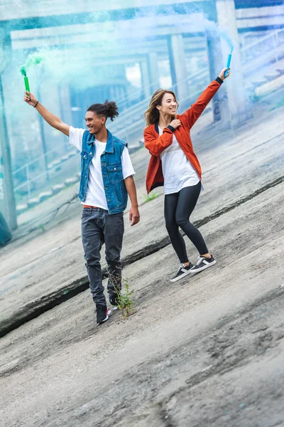 Jeune couple multiculturel tenant des bombes fumigènes colorées dans la rue de la ville — Photo de stock
