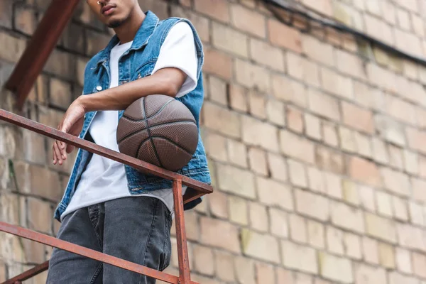 Imagen recortada de hombre joven y elegante de pie con pelota de baloncesto en la calle - foto de stock