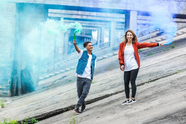 Sorrindo casal multicultural segurando bombas de fumaça coloridas na rua urbana — Fotografia de Stock