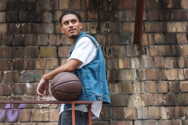 Elegante hombre de raza mixta de pie con pelota de baloncesto y mirando hacia la calle - foto de stock