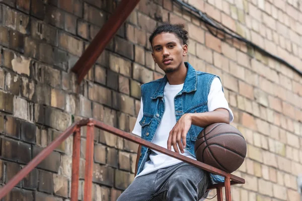 Joven hombre de raza mixta de pie con pelota de baloncesto y mirando a la cámara en la calle - foto de stock