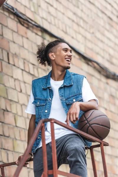 Feliz joven mixto raza hombre de pie con pelota de baloncesto en la calle - foto de stock