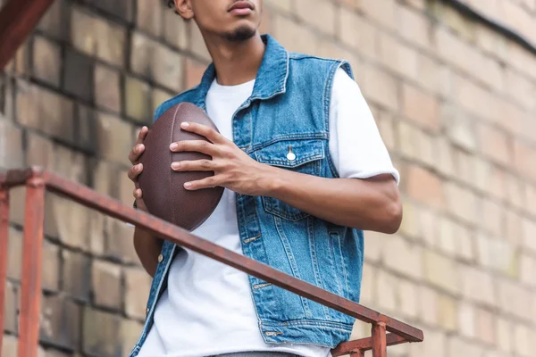 Imagen recortada de un joven sosteniendo pelota de rugby en la calle urbana - foto de stock